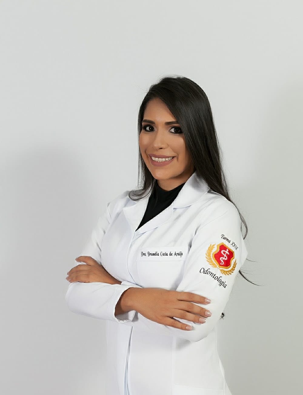 Dra. Brunella Costa de Araújo de jaleco branco, braços cruzados, sorrindo